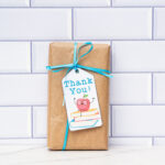 This is a box wrapped in brown kraft paper with a blue ribbon and a gift tag that says thank you with an apple sitting on a stack of books.
