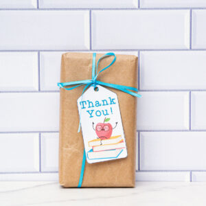 This is a box wrapped in brown kraft paper with a blue ribbon and a gift tag that says thank you with an apple sitting on a stack of books.