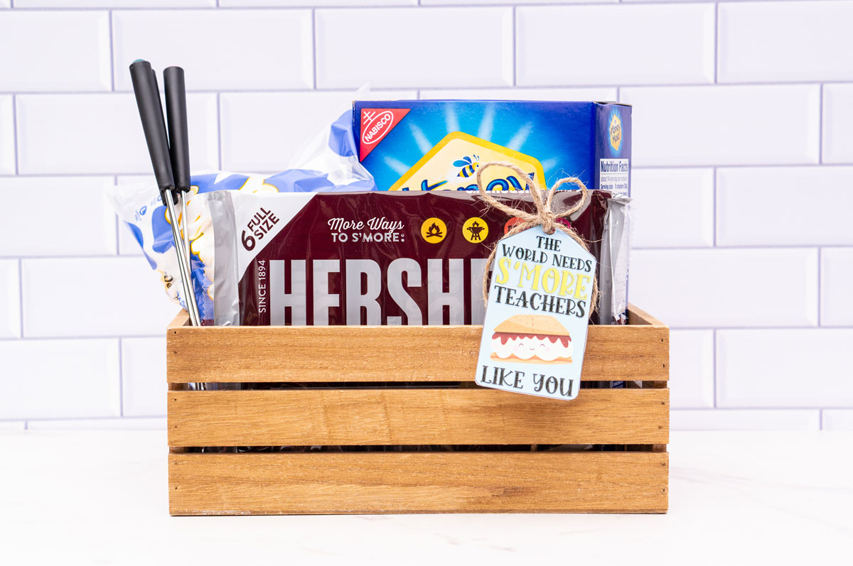 This image shows a wooden crate with chocolate bars, marshmallows, skewers, and a box of graham crackers. The tag says the world needs s'more teachers like you.