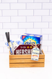 This image shows a wooden crate with chocolate bars, marshmallows, skewers, and a box of graham crackers. The tag says the world needs s'more teachers like you.