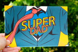 This is a picture of a card that says Super Dad across the chest of a man in a suit open to a superhero costume.
