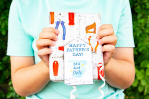 This image shows a boy holding a wrapped present with a gift tag that says Happy Fathers Day with a mug which says best dad ever that you can get from the free Happy Fathers Day tags printable set at the end of this post.