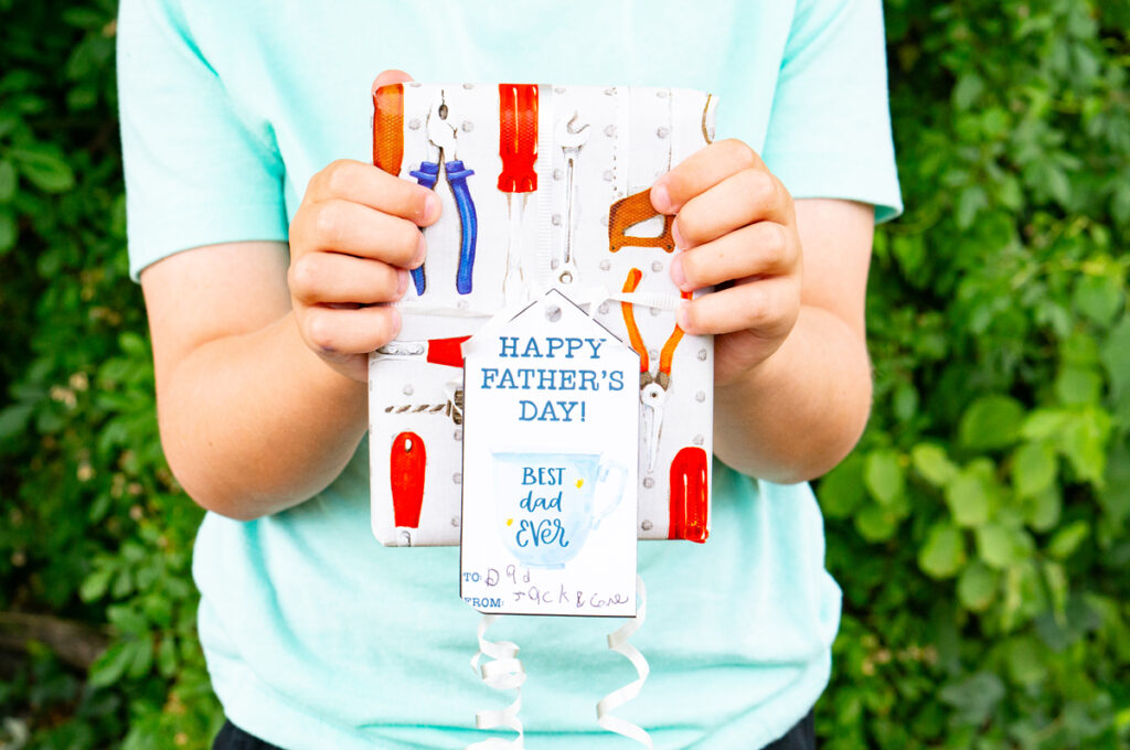 This image shows a boy holding a wrapped present with a gift tag that says Happy Fathers Day with a mug which says best dad ever that you can get from the free Happy Fathers Day tags printable set at the end of this post.