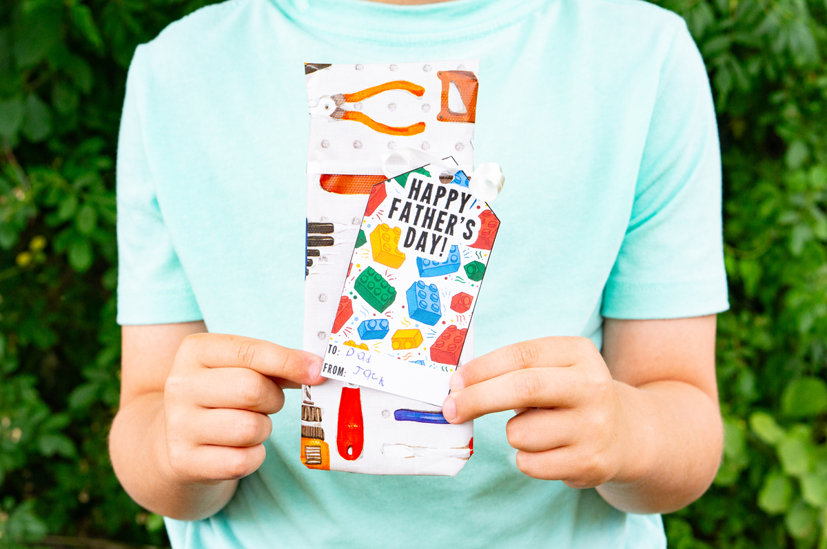 This image shows a boy holding a wrapped present with a gift tag that says Happy Fathers Day with legos that you can get from the free Happy Fathers Day tags printable set at the end of this post.