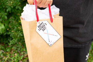 This image shows a boy holding a gift bag with a gift tag that says Happy Fathers Day with 2 light sabers that you can get from the free Happy Fathers Day tags printable set at the end of this post.