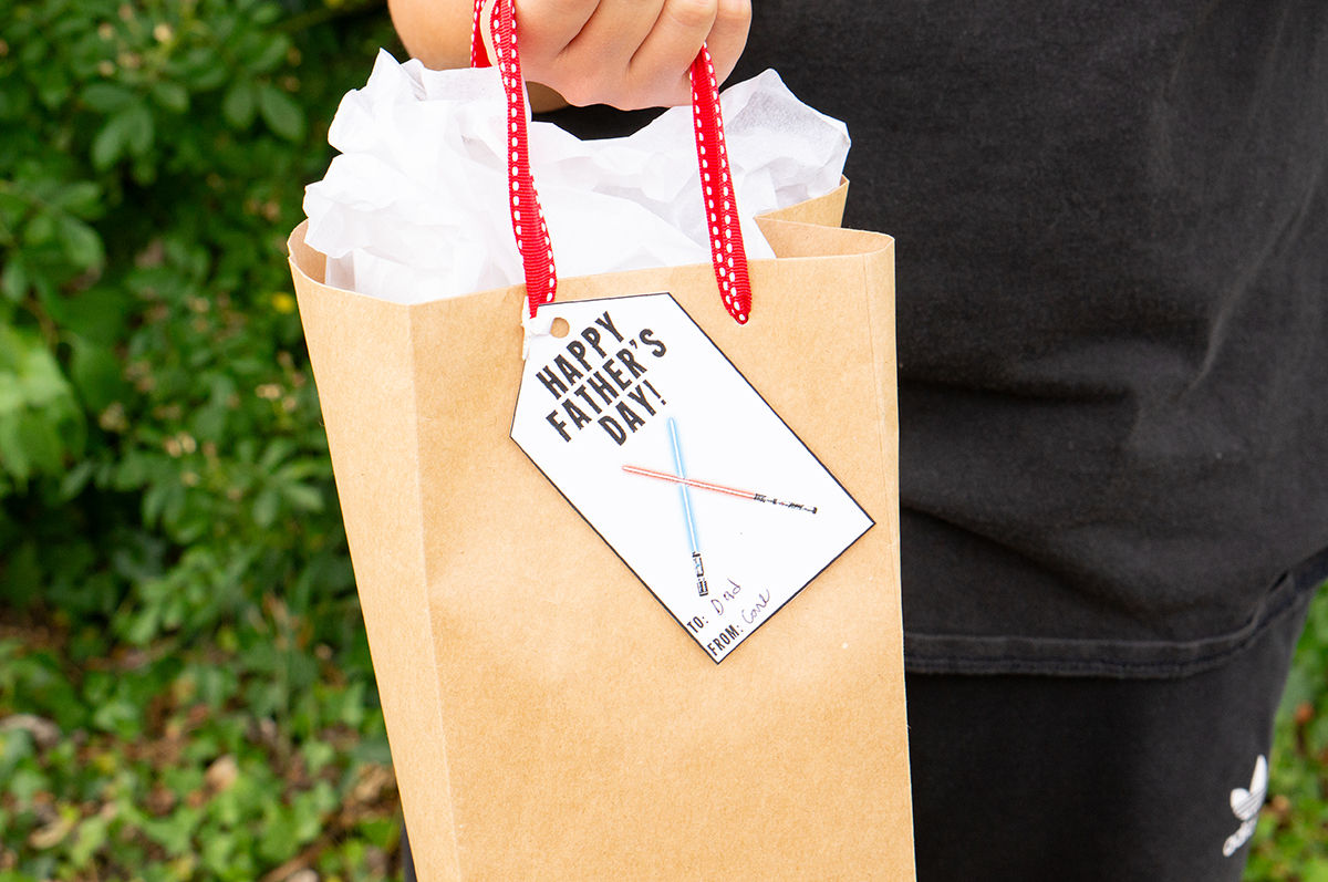 This image shows a boy holding a gift bag with a gift tag that says Happy Fathers Day with 2 light sabers that you can get from the free Happy Fathers Day tags printable set at the end of this post.