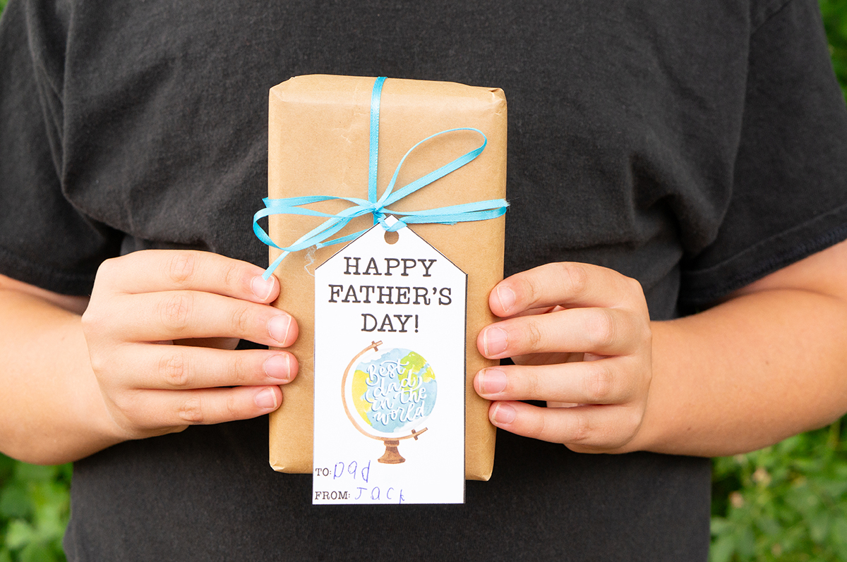 This image shows a boy holding a wrapped present with a gift tag that says Happy Fathers Day with a globe that you can get from the free Happy Fathers Day tags printable set at the end of this post.