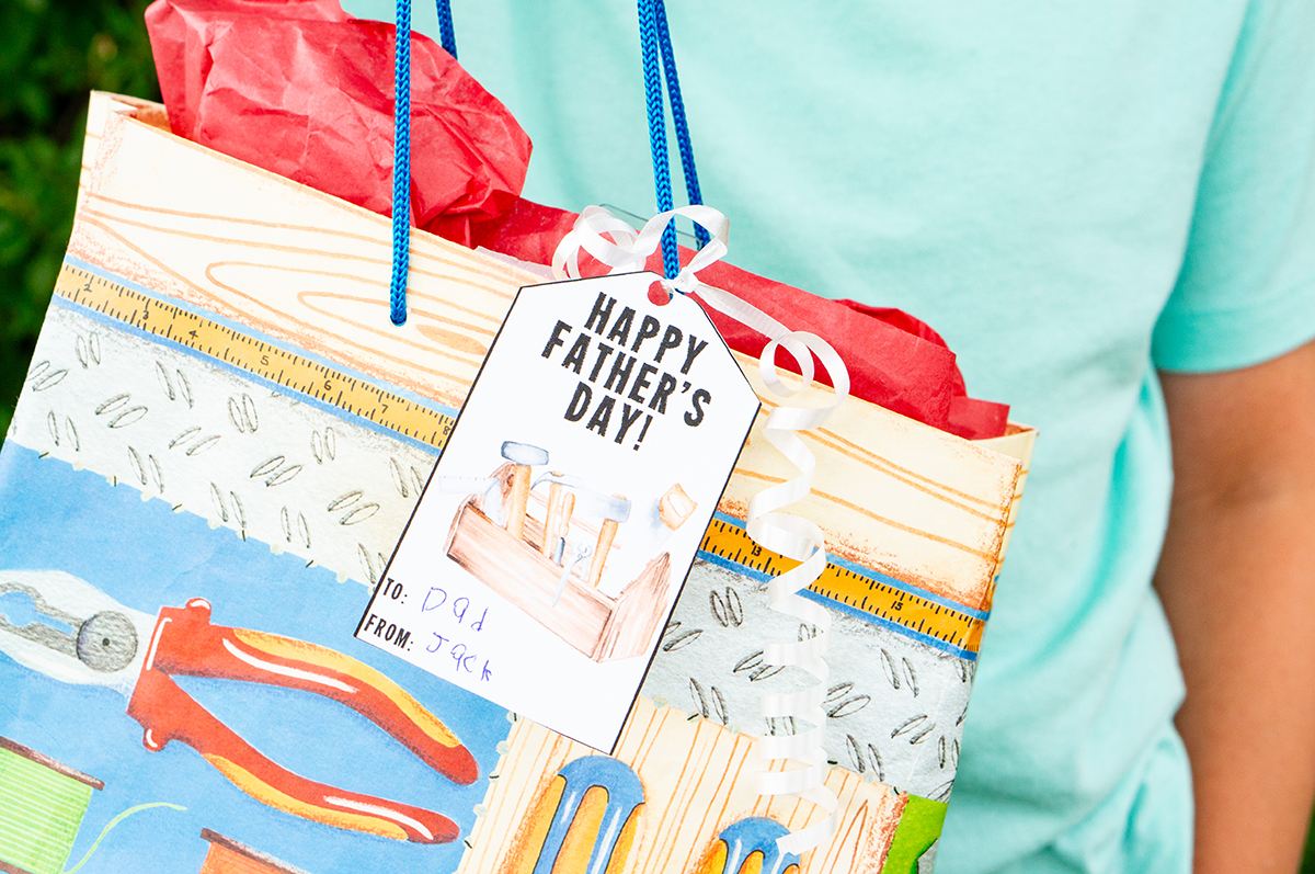 This image shows a boy holding a gift bag with a gift tag that says Happy Fathers Day with a toolbox that you can get from the free Happy Fathers Day tags printable set at the end of this post.