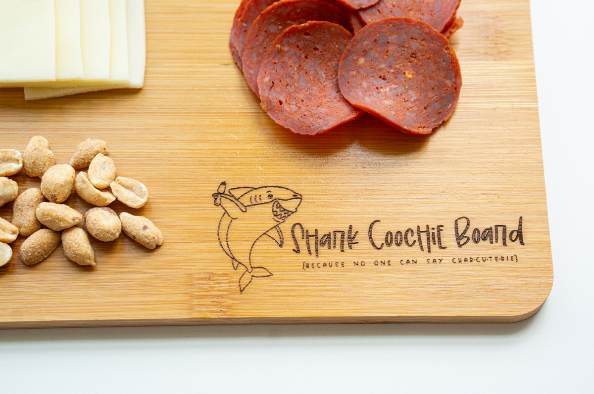 This is an image of a cutting board with the free Shark coochie board SVG engraved on it with some pepperoni, cheese, and peanuts on top of the board.
