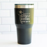 The image is of a laser engraved tumbler that says suck it up buttercup and drink your water - with the drawing of a buttercup flower.