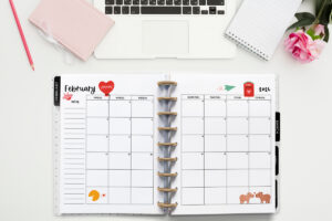 This image is an example of one of the printable 2026 calendar you can get for free at the end of this post. This is showing the month of February inside of an open planner. Above the planner is an open laptop keyboard, some paper, a pencil, headphones, and a flower.