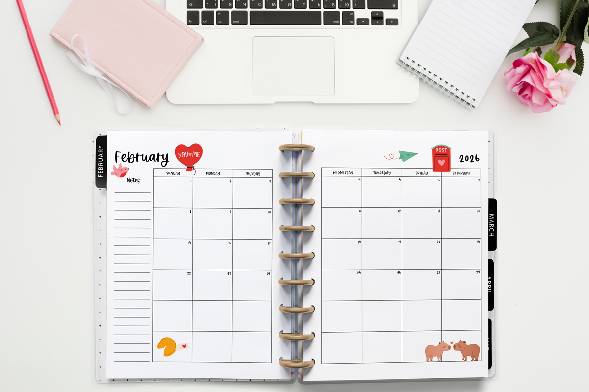 This image is an example of one of the printable 2026 calendar you can get for free at the end of this post. This is showing the month of February inside of an open planner. Above the planner is an open laptop keyboard, some paper, a pencil, headphones, and a flower.
