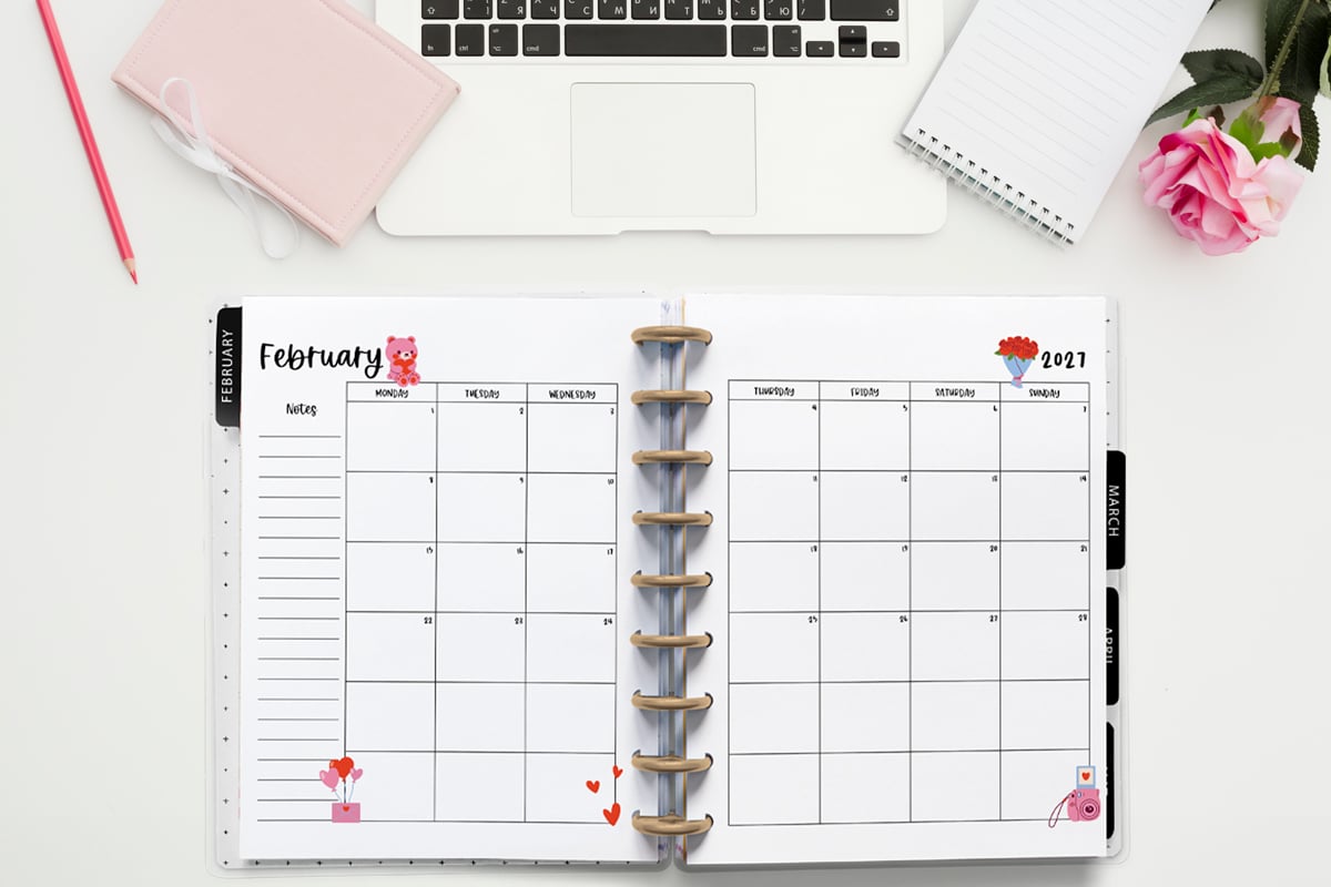 This image is an example of one of the printable 2027 calendar you can get for free at the end of this post. This is showing the month of February inside of an open planner. Above the planner is an open laptop keyboard, some paper, a pencil, headphones, and a flower.