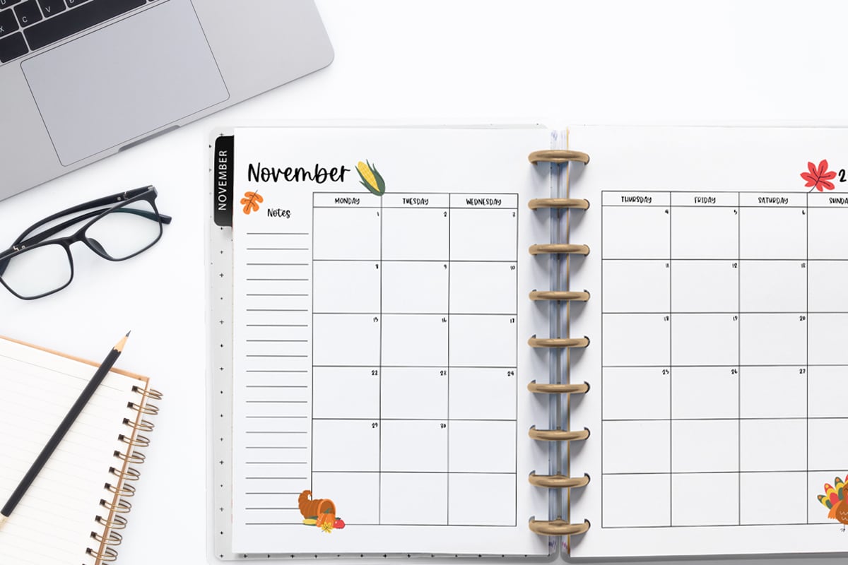 This image is an example of one of the printable 2027 calendar you can get for free at the end of this post. This is showing the month of November inside of an open planner. Above the planner is an open laptop keyboard, some paper, a pencil, and a pair of glasses.