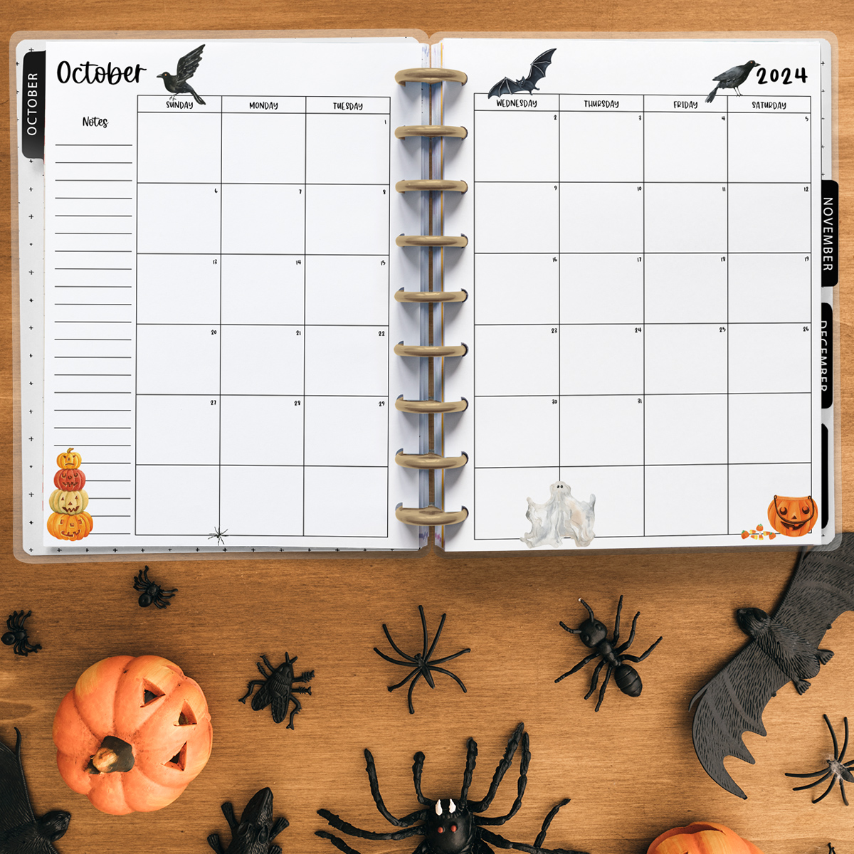 This image is an example of one of the printable 2024 calendar you can get for free at the end of this post. This is showing the month of October inside of an open planner. Below the planner are some fake spiders, bats, and pumpkin.