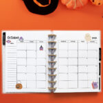 Open planner showing the October 2026 monthly calendar with Halloween decorations. The calendar pages feature a pumpkin, bat, ghost, spell book, spider, and witch illustrations. The background includes pumpkins, a witch hat, a bat cutout, and a black spider against an orange backdrop.