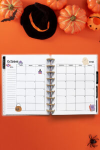 Open planner showing the October 2026 monthly calendar with Halloween decorations. The calendar pages feature a pumpkin, bat, ghost, spell book, spider, and witch illustrations. The background includes pumpkins, a witch hat, a bat cutout, and a black spider against an orange backdrop.