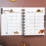 Open planner showing the November 2026 monthly calendar with Thanksgiving-themed illustrations. The calendar features a turkey, pumpkin pie, autumn leaves, and a cornucopia. Surrounding the planner are pumpkins, corn, and seasonal foliage on a brown surface.