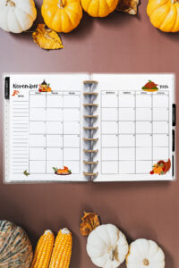 Open planner showing the November 2026 monthly calendar with Thanksgiving-themed illustrations. The calendar features a turkey, pumpkin pie, autumn leaves, and a cornucopia. Surrounding the planner are pumpkins, corn, and seasonal foliage on a brown surface.