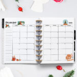 Open planner showing the December 2026 monthly calendar with Christmas decorations. The calendar features illustrations of a gift box, mug of cocoa, ornaments, a snow globe, and holiday cookies. The background includes candy canes, evergreen sprigs, a wooden angel ornament, pinecones, and festive berries.