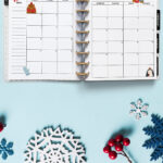 Open planner showing the January 2026 monthly calendar with winter-themed illustrations. The calendar includes a bear, mitten, penguin, and cozy mug drawings. The background features snowflake cutouts, red berries, and a light blue surface for a frosty winter look.