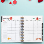Open planner displaying the February 2026 monthly calendar with Valentine’s Day themed decorations, including hearts, candles, and small red embellishments around the planner. The calendar pages feature a red heart with “You + Me,” a red mailbox with a love letter, and two cartoon animals facing each other.