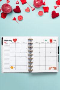Open planner displaying the February 2026 monthly calendar with Valentine&rsquo;s Day themed decorations, including hearts, candles, and small red embellishments around the planner. The calendar pages feature a red heart with &ldquo;You + Me,&rdquo; a red mailbox with a love letter, and two cartoon animals facing each other.
