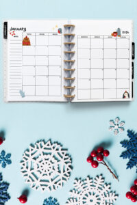 Open planner showing the January 2026 monthly calendar with winter-themed illustrations. The calendar includes a bear, mitten, penguin, and cozy mug drawings. The background features snowflake cutouts, red berries, and a light blue surface for a frosty winter look.
