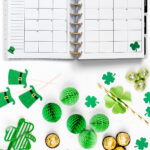 Open planner showing the March 2026 monthly calendar with St. Patrick’s Day decorations, surrounded by green shamrocks, leprechaun hats, and gold-wrapped candies. The calendar pages feature a green pipe, pot of gold, shamrock, and leprechaun hat illustrations.