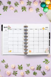 Open planner showing the April 2026 monthly calendar with Easter and spring-themed decorations. The calendar includes small illustrations of chicks, flowers, and a bunny. Around the planner are soft pink roses, greenery, and colorful dyed Easter eggs.