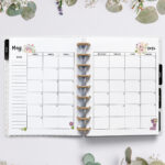 Open planner showing the May 2026 monthly calendar with floral and gardening illustrations. The calendar has flower pots and blooms decorating the corners. The surrounding background features fresh greenery, daisies, lavender, and delicate spring flowers.