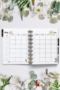 Open planner showing the May 2026 monthly calendar with floral and gardening illustrations. The calendar has flower pots and blooms decorating the corners. The surrounding background features fresh greenery, daisies, lavender, and delicate spring flowers.