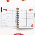 Open planner showing the June 2026 monthly calendar with strawberry-themed decorations. The calendar pages feature strawberry and cake illustrations. Fresh strawberries are placed above and below the planner on a clean white background.