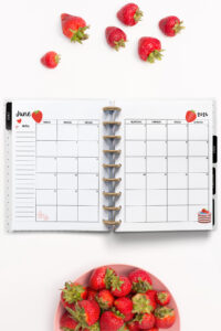 Open planner showing the June 2026 monthly calendar with strawberry-themed decorations. The calendar pages feature strawberry and cake illustrations. Fresh strawberries are placed above and below the planner on a clean white background.