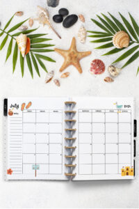 Open planner showing the July 2026 monthly calendar with beach and summer-themed decorations. The calendar includes flip-flops, a seagull, a beach sign, and a sandcastle illustration. The surrounding background has seashells, a starfish, palm leaves, and smooth stones on white sand.