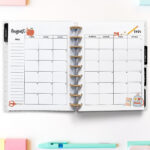 Open planner showing the August 2026 monthly calendar with back-to-school themed decorations. The calendar pages feature a red apple with a worm, scissors, a pencil, a notebook, and a backpack illustration. Surrounding the planner are colorful stationery supplies, including sticky notes, pencils, pens, washi tape, and pastel push pins.