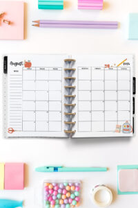 Open planner showing the August 2026 monthly calendar with back-to-school themed decorations. The calendar pages feature a red apple with a worm, scissors, a pencil, a notebook, and a backpack illustration. Surrounding the planner are colorful stationery supplies, including sticky notes, pencils, pens, washi tape, and pastel push pins.