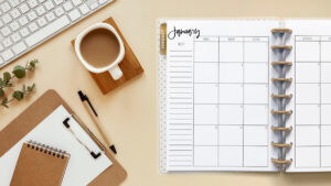 This image is an example of one of the printable 2024 calendar you can get for free at the end of this post. This is showing the month of January without holidays or clipart inside of an open planner. Next to the planner is a cup of coffee, clipboard with paper, a small notebook, and part of a computer keyboard.