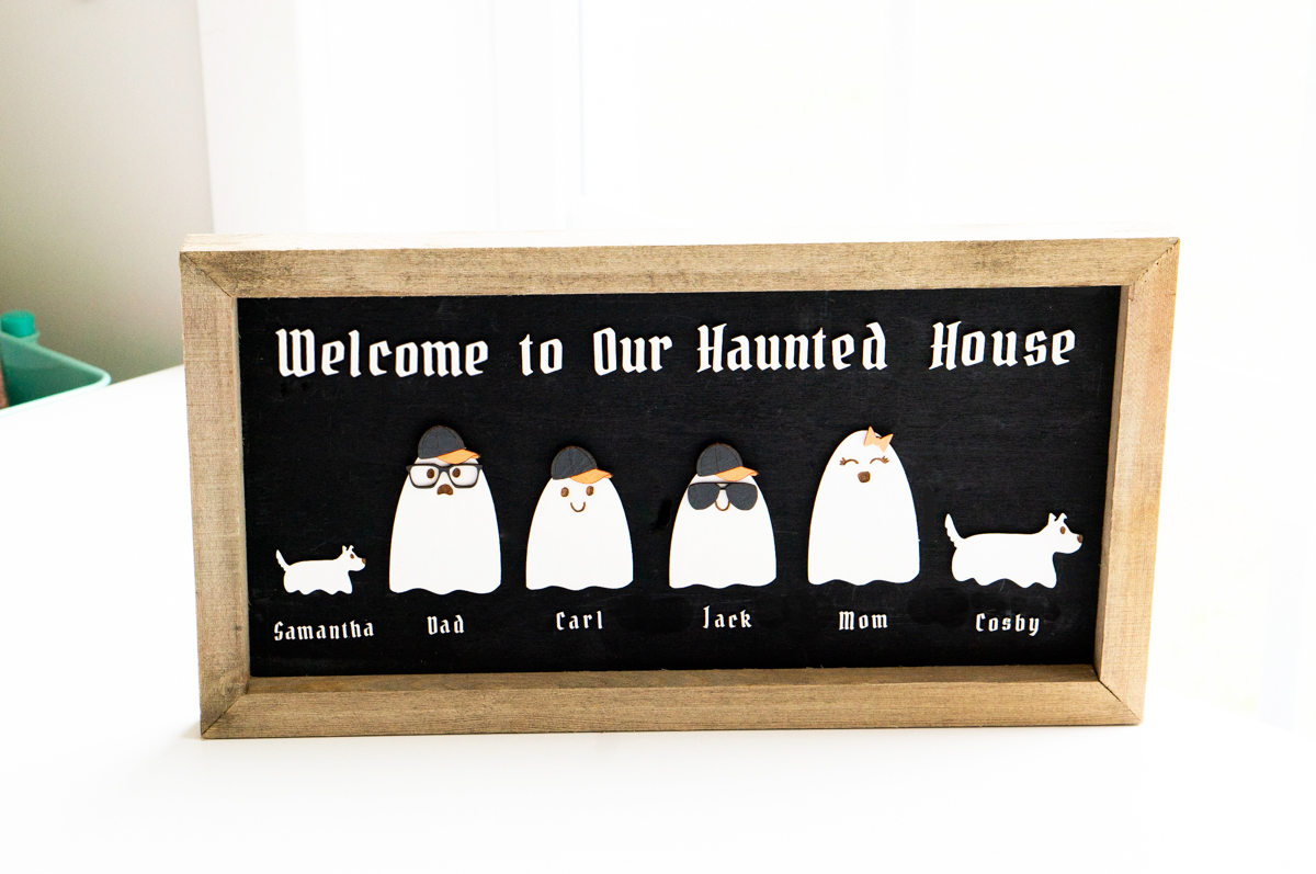 This image shows the completed example of the Halloween ghost family wood sign using the free ghost family SVG set. At the top it says welcome to our haunted house. Then there are some ghosts in the middle with the names, Samantha, Dad, Carl, Jack, Mom, and Cosby.