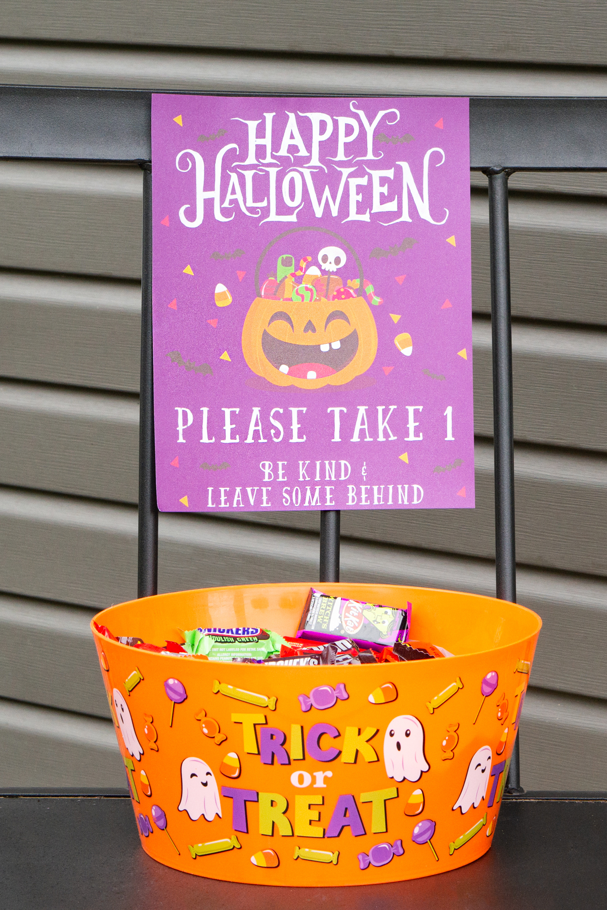 This image shows one of the free printable Halloween candy signs you can get in this free set. This sign says Happy Halloween. Please take 1. Be kind and leave some behind. Under the sign is a bowl of candy.