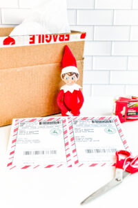 This image shows an example of the free Elf on the shelf mailing label you can download for free in this blog post. It has a picture of a box, an elf, scissors, packing tape, and 2 labels.