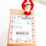 This image shows an example of the free Elf on the shelf mailing label you can download for free in this blog post on a shipping box with an Elf on the Shelf doll escaping out of the top of the box.
