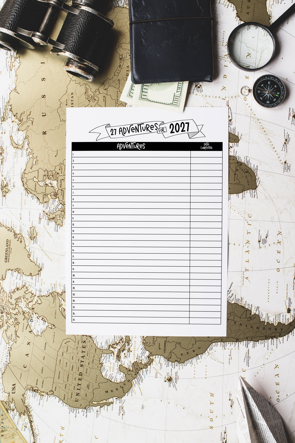 Adventures for 2027 printable sheet laid on top of a vintage world map with travel accessories including binoculars, a wallet, cash, and a magnifying glass.