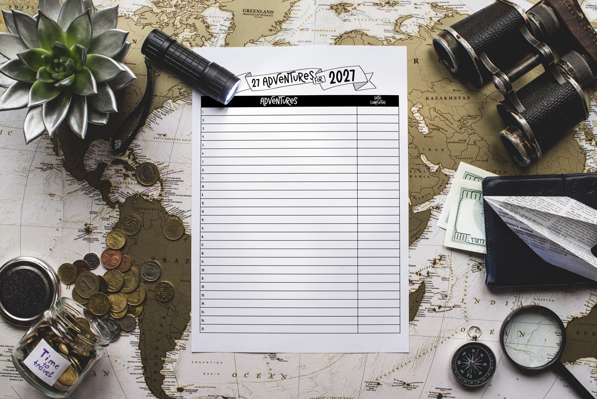 Adventures for 2027 printable placed on a world map surrounded by travel-themed props such as coins, a flashlight, a succulent plant, binoculars, and a compass.
