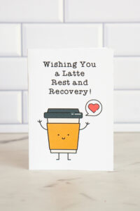 A "Get Well Soon" card featuring an illustrated coffee cup with a smiling face, holding a speech bubble with a heart. The text reads "Wishing You a Latte Rest and Recovery!" blending humor and care.