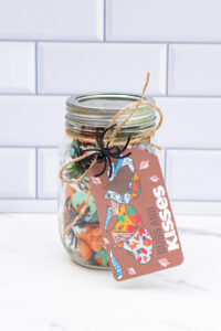 This is an image of a mason jar filled with Hershey Kisses and fake plastic spiders. It has a free printable Halloween tag hanging from the jar that says: Bugs and Kisses.