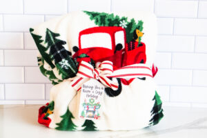 This is one of the free Christmas treat gift tags you can download in this post. It says, Wishing You a Warm & Cozy Holiday. It features a fireplace design with holiday decor, a wreath overtop, and Christmas decor. The tag is attached to a blanket with red Christmas trucks on it.