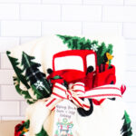 This is one of the free Christmas treat gift tags you can download in this post. It says, Wishing You a Warm & Cozy Holiday. It features a fireplace design with holiday decor, a wreath overtop, and Christmas decor. The tag is attached to a blanket with red Christmas trucks on it.