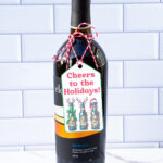 This is one of the free Christmas treat gift tags you can download in this post. It says, Cheers to the Holidays. It features bottles of beer decorated with Christmas lights, antlers, and one has a Santa hat. The tag is attached to a bottle of wine.