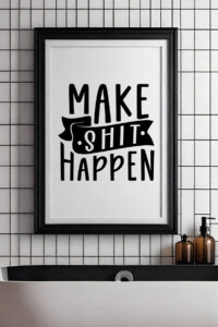 Framed bathroom sign featuring the bold and humorous phrase 'Make Shit Happen' in black typography. The sign is mounted on a sleek white tiled wall above a modern black and white freestanding sink, with amber soap dispensers adding a touch of warmth to the minimalist decor.